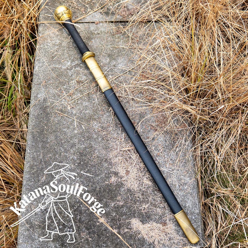May include: A black walking stick with gold-colored accents and a rounded top. The stick has a gold-colored band near the top and bottom. The text "Katana SoulForge" is visible in the lower left corner.