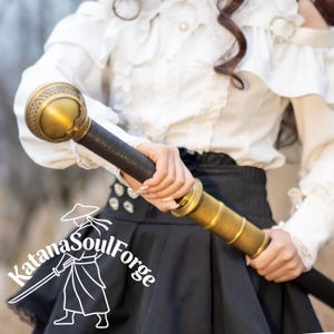 May include: A decorative sword with a gold-colored hilt and accents, a black handle, and a black blade. The sword is held in front of a person wearing a white blouse and black skirt. The Katana SoulForge logo is in the lower left corner.