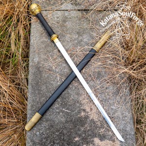 Peut inclure: Une épée avec une lame argentée et une poignée noire, posée sur une surface en pierre. La poignée a un pommeau et des accents dorés. Le fourreau est noir avec des accents dorés. Le texte "Katana SoulForge" est visible.