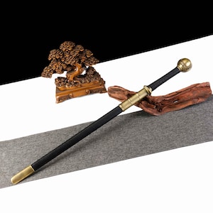 Pode incluir: Uma espada preta e dourada com um pomo redondo e uma lâmina longa e reta. A espada tem uma empunhadura preta e detalhes dourados. A espada é exibida em uma superfície cinza com uma escultura de madeira ao fundo.