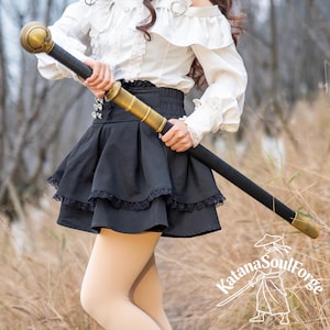 May include: A black and gold staff with a decorative top and bottom. The staff is held by a person wearing a white blouse and black skirt. The staff has a black handle and gold accents. The Katana SoulForge logo is visible in the bottom right corner.