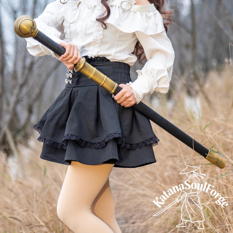 May include: A black and gold staff with a decorative top and bottom. The staff is held by a person wearing a white blouse and black skirt. The staff has a black handle and gold accents. The Katana SoulForge logo is visible in the bottom right corner.