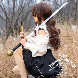 May include: A long, silver katana sword with a black handle and gold accents. The sword is held by a person wearing a white blouse and black skirt. The Katana SoulForge logo is visible on the skirt.