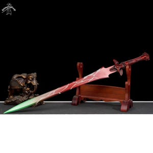Peut inclure: Une épée décorative rouge et blanche avec une pointe verte, présentée sur un support en bois. L'épée présente une poignée et une lame ornées. Une statue d'éléphant en bronze est visible en arrière-plan. C'est un objet décoratif.