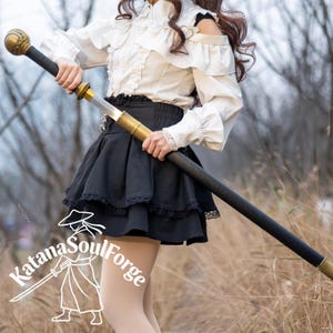 May include: A black and gold staff with a decorative top and bottom. The staff is held by a person wearing a white blouse and black skirt. The text "Katana SoulForge" is visible in the lower left corner.