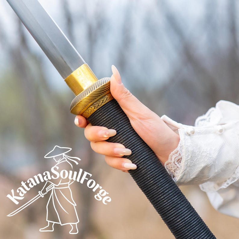 May include: A close-up of a katana sword. The sword features a black handle with a textured grip, a gold-colored handguard, and a silver blade. The Katana SoulForge logo is visible in the lower left corner.