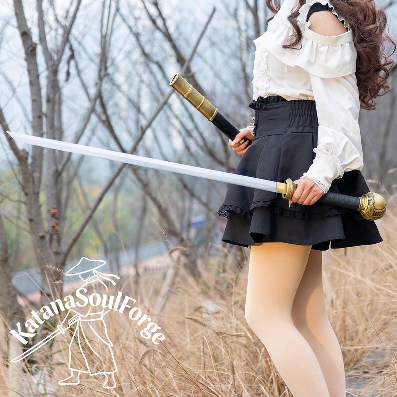 May include: A katana with a gold and black handle and a silver blade. The sword is held in front of a person wearing a white blouse and black skirt. The Katana SoulForge logo is in the bottom left corner.