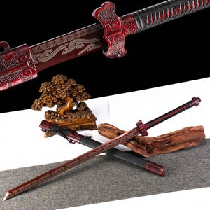 May include: A red and silver sword with intricate designs, featuring a decorative hilt and blade. The sword is displayed with a black sheath and a wooden sculpture. The blade has a silver pattern.