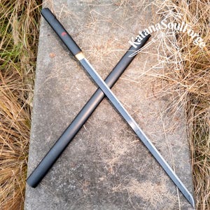 May include: A black katana sword with a silver blade, resting on a stone surface. The sword's scabbard is black with a red accent. The text "Katana SoulForge" is visible on the scabbard.