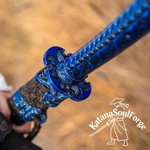 Pode incluir: Close-up de uma espada katana azul e preta. A espada apresenta intrincados padrões florais e ondulados na empunhadura e na guarda. A lâmina não é visível. O logotipo Katana Soul Forge está no canto inferior direito.