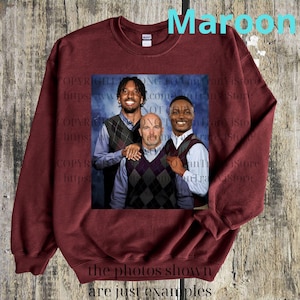 May include: Maroon sweatshirt with a framed image of three men wearing argyle vests over button-down shirts. The text "Maroon" is at the top, and "the photos shown are just examples" is at the bottom. The background is a wooden surface.