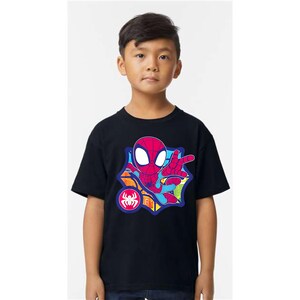 K&ouml;nnte beinhalten: Ein schwarzes T-Shirt mit einer farbenfrohen Cartoon-Grafik von Spider-Man. Das Design zeigt eine rote Spider-Man-Figur mit wei&szlig;en Augen, einem blau-roten Anzug und einem Spinnenemblem. Das T-Shirt wird von einer jungen Person getragen.