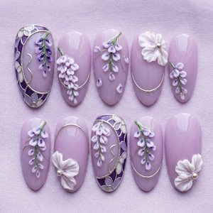 May include: A set of ten almond-shaped press-on nails in a light lavender base color. The designs feature 3D sculpted wisteria flowers in shades of purple and white, silver metallic line accents, and white floral relief details with small silver beads.