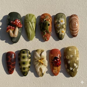 Könnte beinhalten: Ein Set aus zehn mandelförmigen Press-on-Nägeln mit naturinspirierten Designs, darunter rot-weiße Pilze, ein grüner Frosch, weiße Blumen, ein Marienkäfer, eine weiße Lilie sowie verschiedene erdige Muster in Grün-, Braun- und Orangetönen.