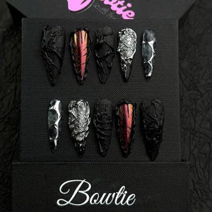 May include: A set of ten press-on nails arranged in two rows on a black textured display box. The nails feature varied designs including matte black with raised vein textures, metallic red-orange chrome, silver scales, and black marble patterns.