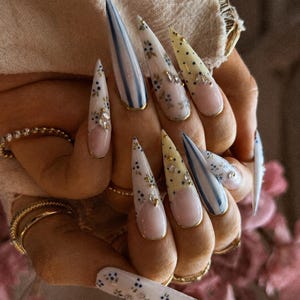 Op de afbeelding: Stiletto-vormige press-on nagels met een mix van ontwerpen, waaronder blauwe verticale strepen, gele stippen en delicate blauwe bloemmotieven op een witte basis. Verschillende nagels zijn versierd met kleine doorzichtige strasssteentjes en goudkleurige randen.