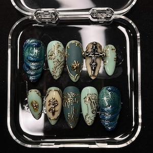 May include: A set of ten artificial nails in a clear plastic case. The nails feature various designs, including gold embellishments, crosses, and swirling patterns in shades of blue, green, and cream.