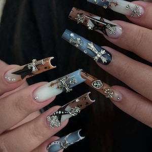 Puede incluir: Uñas largas y esculpidas con diversos diseños. Algunas uñas presentan un degradado azul y blanco, otras un patrón marrón y negro. Cada uña está adornada con dijes plateados de cruces y estrellas.