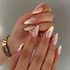 May include: Long almond-shaped nails featuring a glossy nude base with pale yellow French tips. Several nails are adorned with a raised, textured shell-like pattern and small silver studs. A gold-toned metal watch and a gold ring are visible.