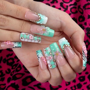 May include: Long square-shaped artificial nails featuring a mix of mint green and nude tones. The designs include 3D pink floral accents, gold rhinestones, white honeycomb patterns, and black animal print details on a glossy, smooth finish.