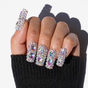 May include: A set of press-on nails featuring a square shape and a full coverage design. Each nail is densely encrusted with iridescent, multi-faceted rhinestones in various shapes, including hearts and butterflies, creating a high-shine finish.