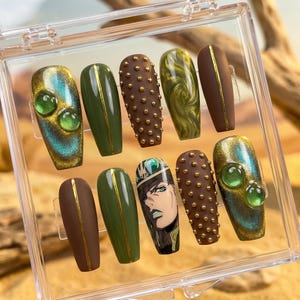 Könnte beinhalten: Ein Set aus zehn künstlichen Nägeln in einer transparenten Kunststoffbox. Die Nägel weisen verschiedene Designs auf, darunter Olivgrün, Braun und Türkis mit goldenen Akzenten. Einige Nägel haben eine Charakterillustration, andere dekorative Nieten oder Edelsteine.