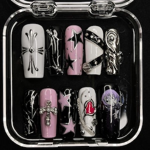 Könnte beinhalten: Ein Set aus zehn künstlichen Nägeln in einer transparenten Kunststoffbox. Die Nägel weisen verschiedene Designs auf, darunter Kreuze, Sterne und abstrakte Muster. Farben sind Weiß, Schwarz, Rosa und Silber.