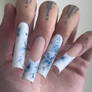 May include: Long, square-shaped artificial nails with a white base and a blue floral design. The nails feature a delicate watercolour-style pattern of blue roses and leaves. The nail beds are a natural pink colour.