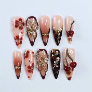 May include: A collection of ten long, almond-shaped artificial nails. The nails feature various designs, including floral patterns, gold accents, and cherry embellishments. Colors include shades of pink, brown, and red, with some nails showcasing a leopard print.