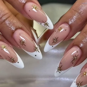 Peut inclure: Ongles en forme d'amande arborant une manucure française classique avec des pointes blanches sur une base nude. Chaque ongle est orné de petits accents dorés en forme d'étoile et de minuscules clous sombres disposés sur la surface.