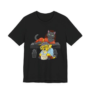 Può includere: T-shirt nero con un'illustrazione di cartoni animati. Un gatto grigio in una veste arancione è sdraiato sopra un cane giallo con occhiali e una camicia blu, che tiene un barattolo di snack. Il design include piccole statue grigie.