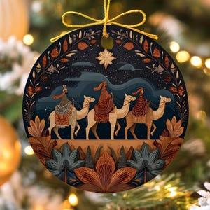 Könnte beinhalten: Runder Christbaumschmuck mit goldenem Band. Der Anhänger zeigt eine Illustration von drei Figuren auf Kamelen unter einem Sternenhimmel. Das Design verwendet Braun-, Blau- und Goldtöne mit floralen Elementen.