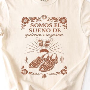 May include: Cream-colored t-shirt with a brown graphic. The graphic includes the text "SOMOS EL SUENO DE quienes cruzaron" above a butterfly, sun, and sandals design. Floral and cactus elements frame the design.