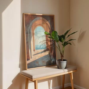 May include: A framed artwork depicting a series of archways leading to a seascape, with a light brown wooden frame. The artwork is displayed on a light beige bench with a potted plant. The scene is bathed in sunlight, creating shadows.