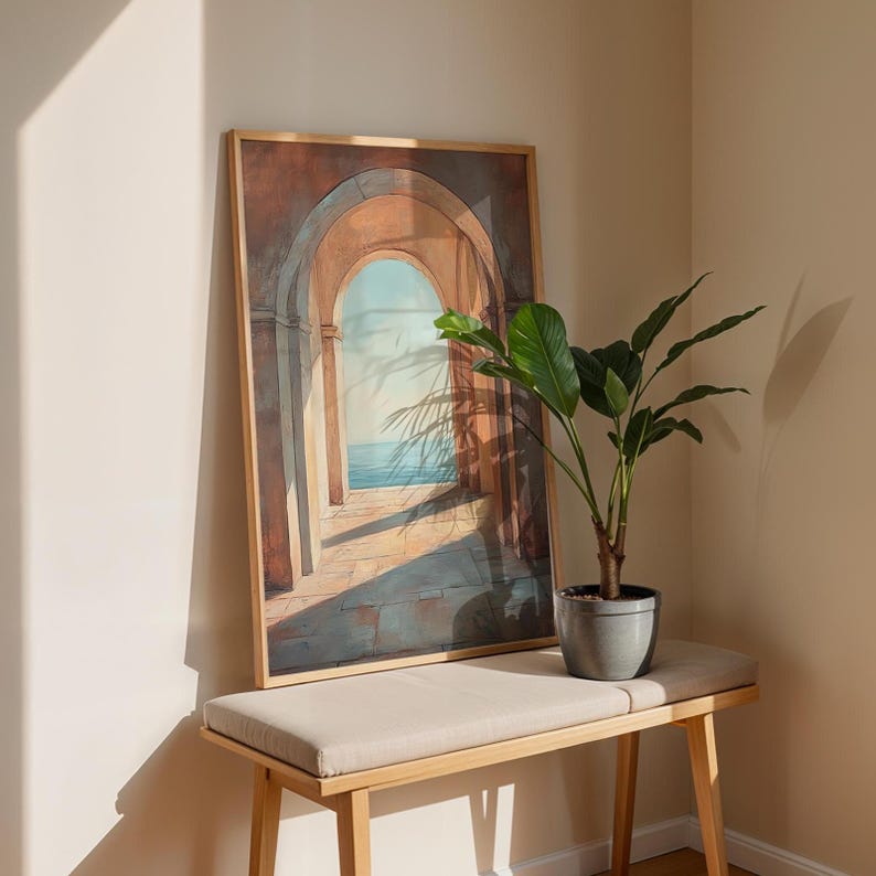 May include: A framed artwork depicting a series of archways leading to a seascape, with a light brown wooden frame. The artwork is displayed on a light beige bench with a potted plant. The scene is bathed in sunlight, creating shadows.