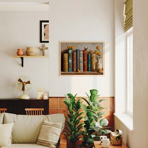 May include: A framed print featuring a still life of antique books with colorful spines, a butterfly, and yellow flowers in a vase. The artwork is displayed on a white wall, with a shelf and other decorative items visible.