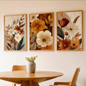 Könnte beinhalten: Drei gerahmte Kunstdrucke mit Blumenmotiven. Jeder Druck zeigt Blumen im Aquarellstil in Creme-, Orange- und Brauntönen. Das Kunstwerk ist vor einem neutralen Hintergrund platziert, und die Rahmen sind hellbraun. Das Gesamtthema ist botanisch.