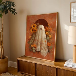 May include: A framed art print featuring a ghost figure draped in a floral lace sheet, set against a rust-colored backdrop. The ghost is surrounded by sunflowers and other autumnal flowers. The artwork has a gold frame and is displayed on a wooden cabinet.