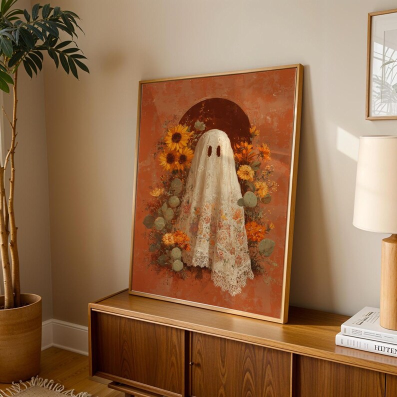 May include: A framed art print featuring a ghost figure draped in a floral lace sheet, set against a rust-colored backdrop. The ghost is surrounded by sunflowers and other autumnal flowers. The artwork has a gold frame and is displayed on a wooden cabinet.