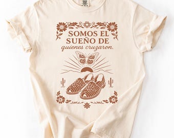 May include: Cream-colored t-shirt with a brown graphic design. The design includes the text "SOMOS EL SUEÑO DE quienes cruzaron." A butterfly, sandals, sun, and cacti are also featured in the design. The shirt has rolled-up sleeves.