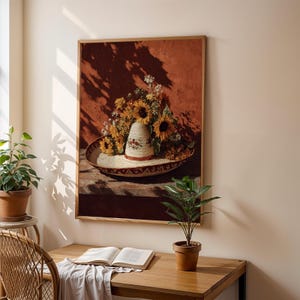 May include: A framed still life painting depicts a sombrero filled with sunflowers and other flowers. The sombrero sits in a decorative bowl on a wooden surface against a reddish-brown wall. The painting is illuminated by sunlight.