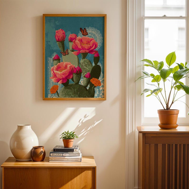 May include: A framed art print featuring a cactus with large pink flowers and butterflies against a blue background. The artwork is in a wooden frame and is displayed on a light-colored wall. A small potted plant and decorative vases are on a wooden cabinet below.