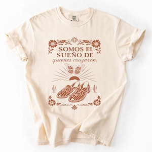 May include: A cream-colored short-sleeve t-shirt featuring a rust-colored graphic print. The design includes the text "SOMOS EL SUEÑO DE quienes cruzaron." above a butterfly, a rising sun, and a pair of huarache sandals, framed by floral motifs.