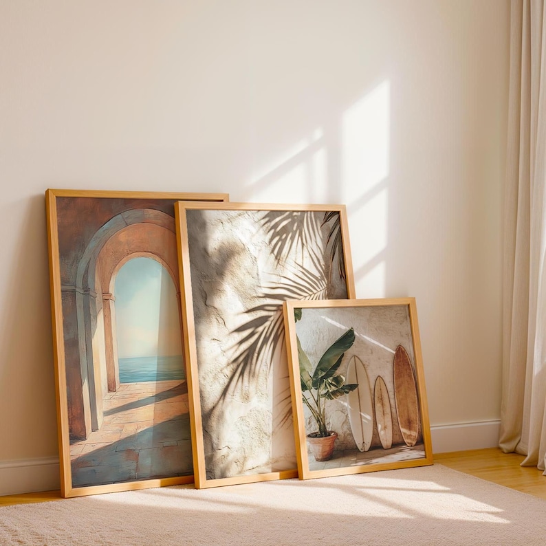 May include: Three framed art prints leaning against a wall. The prints feature images of an archway overlooking the sea, palm fronds, and surfboards with a potted plant. The frames are made of light-colored wood.
