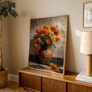 May include: A framed painting depicts a still life of orange marigold flowers in a brown vase. The vase sits on a surface with blue and green tiles. The painting has a gold frame and is displayed on a wooden cabinet.