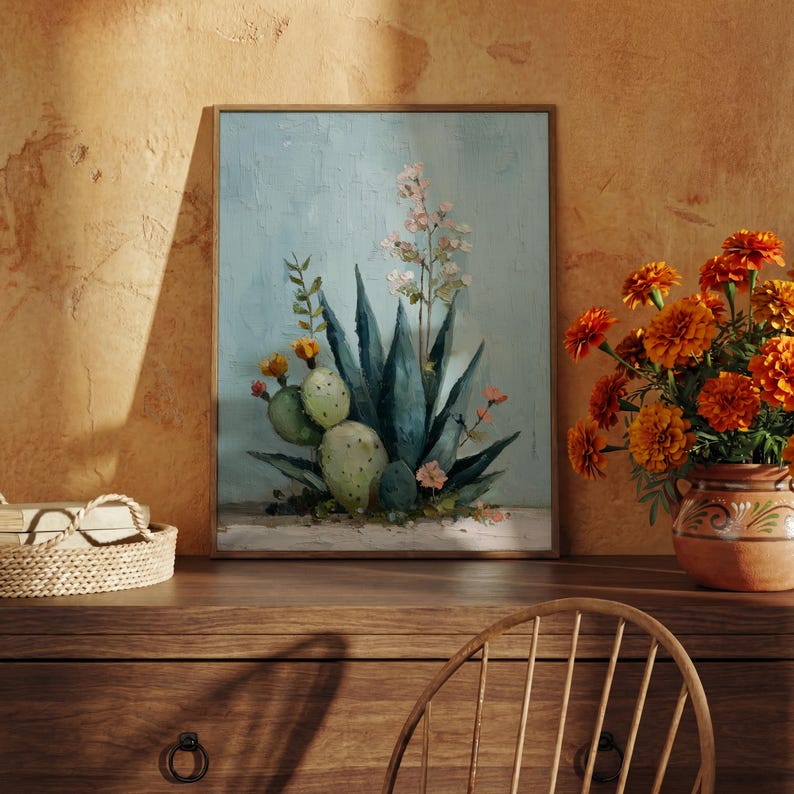 May include: A framed painting of a desert scene featuring a blue agave plant, cacti, and delicate flowers. The artwork is set against a light blue background and framed in a wooden frame. A vase of orange flowers sits nearby.