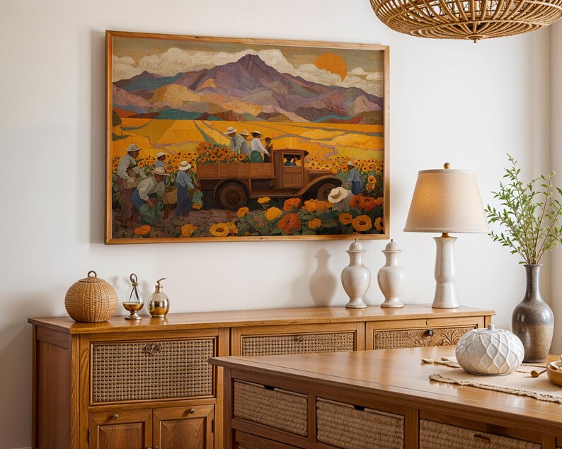 May include: A framed painting depicts a field of sunflowers with workers harvesting them. A vintage truck is loaded with sunflowers. The background features mountains and a setting sun. The painting is in a wooden frame and hangs above a wooden cabinet.