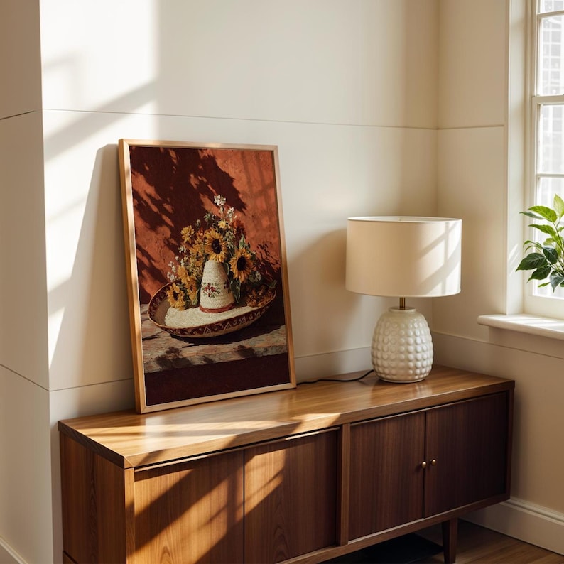 May include: A framed artwork featuring a still life of sunflowers and a sombrero. The artwork is in a wooden frame and rests on a wooden cabinet. A white lamp with a textured base sits on the cabinet, and a window is visible in the background.
