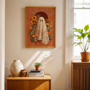 May include: A framed art print featuring a ghost figure draped in a floral lace cloth. The artwork is set against a terracotta background with sunflowers and other flowers. The frame is made of wood.