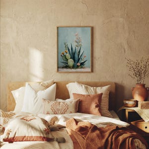 May include: A framed watercolor print of a cactus and other desert plants hangs above a bed with decorative pillows and a rust-colored blanket. The artwork features shades of green, blue, and pink against a light blue background. The bed has a neutral color palette.